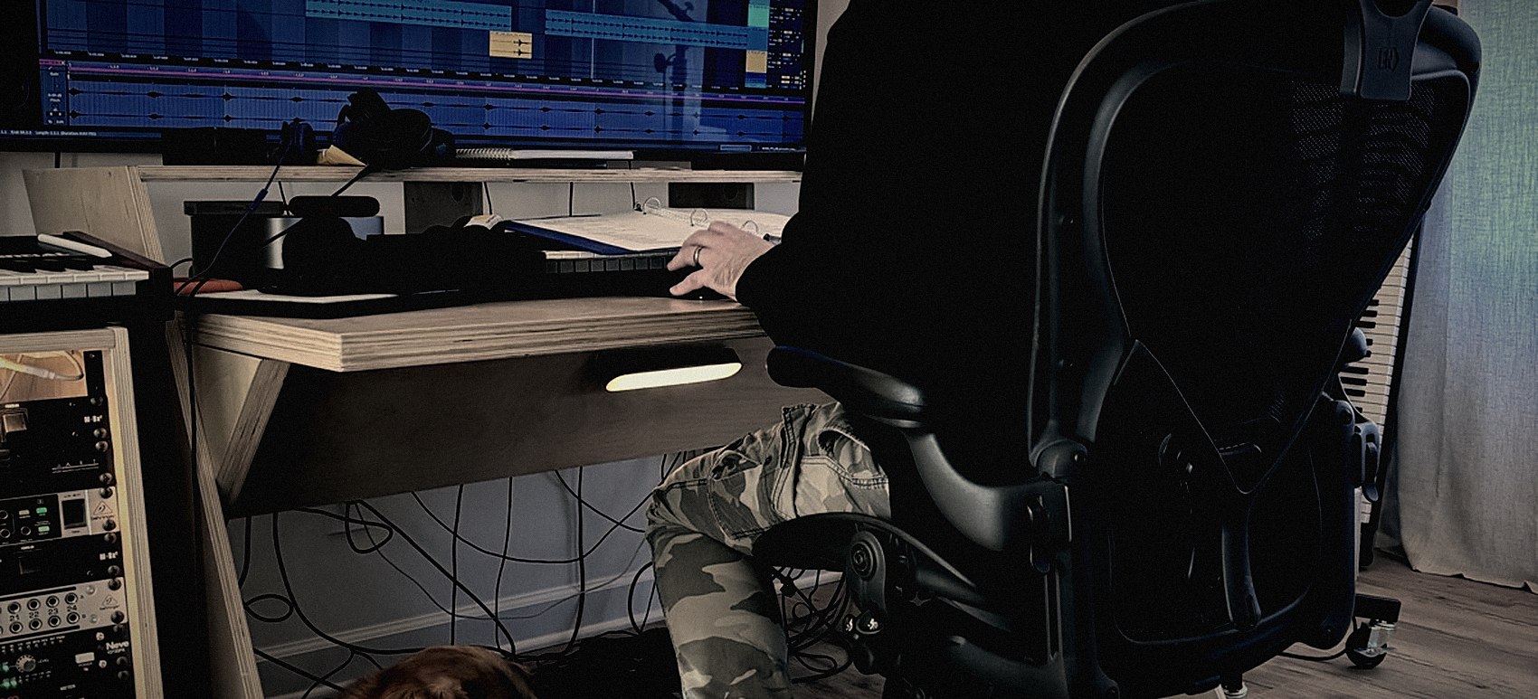 Brock at the studio desk with a session open on the monitor, the dog asleep underneath